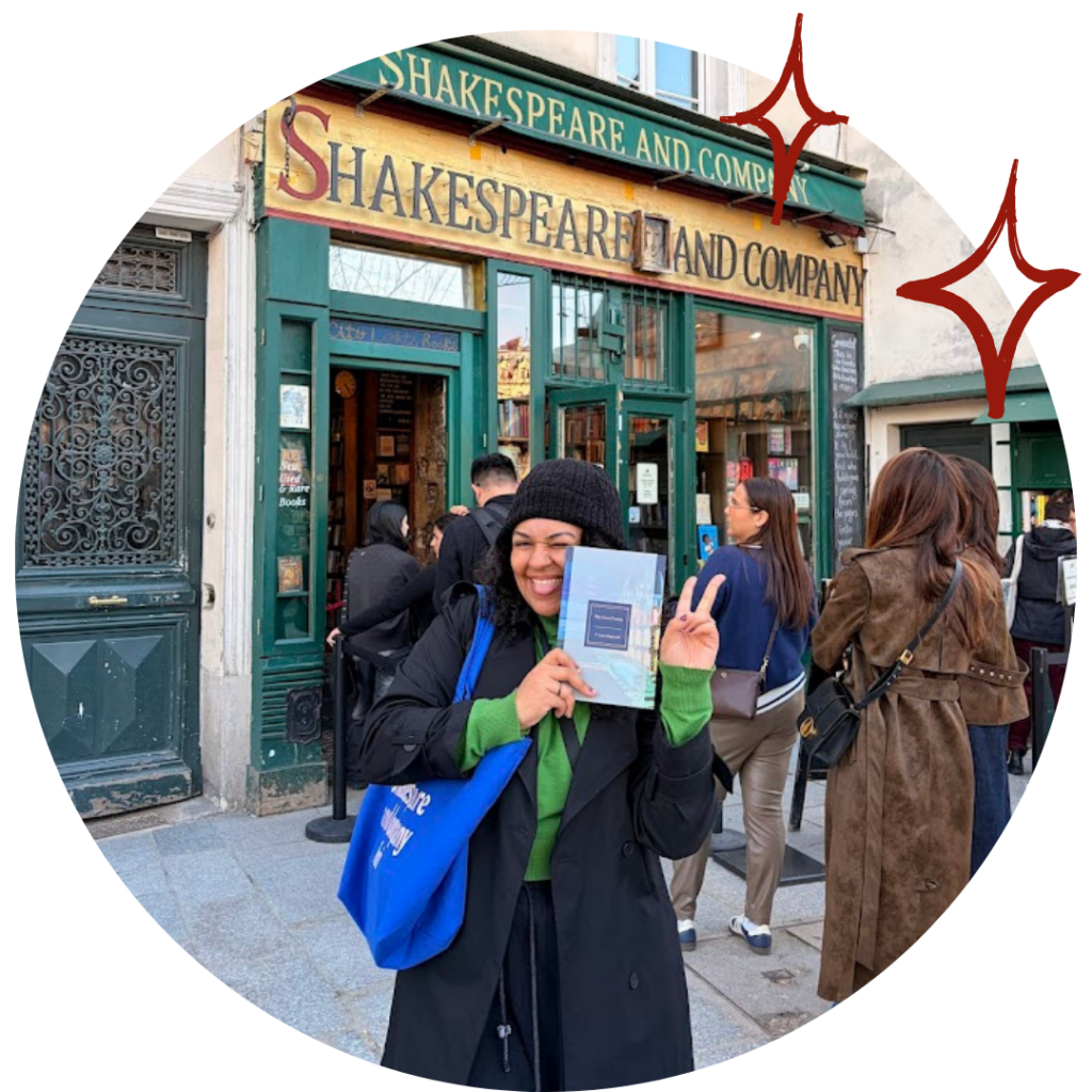 Uma foto de Karoline Melo. Mulher de 26 anos, pele negra de tom marrom-claro. Está no centro da imagem, usando uma touca preta, um sobretudo preto, um suéter verde, uma calça preta e com uma tote bag azul. Está segurando o livro The Great Gatsby com uma mão e fazendo sinal da paz com a outra. Sua expressão está sorridente, com uma careta leve: piscando um olho e mordendo a língua de um jeito divertido. Atrás dela, uma fila de pessoas prontas para entrar na livraria Shakespeare and Company, em Paris. A fachada da livraria é simpes e verde-bandeira, com letreiro amarelo-ovo e detalhes vermelhos. A imagem é redonda, e do lado direito há duas estrelas de quatro pontas desenhadas de vermelho.